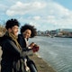 Young Couple of Friends Are meeting in Town for a takeaway Coffee a Walk by the river. The urban area is lighted by sunny cold autumn/ winter day. the Latin Man and the Afro Black Woman are Wearing Heavy jackets and holding a Cup of Coffee from the Shon in The Corner.