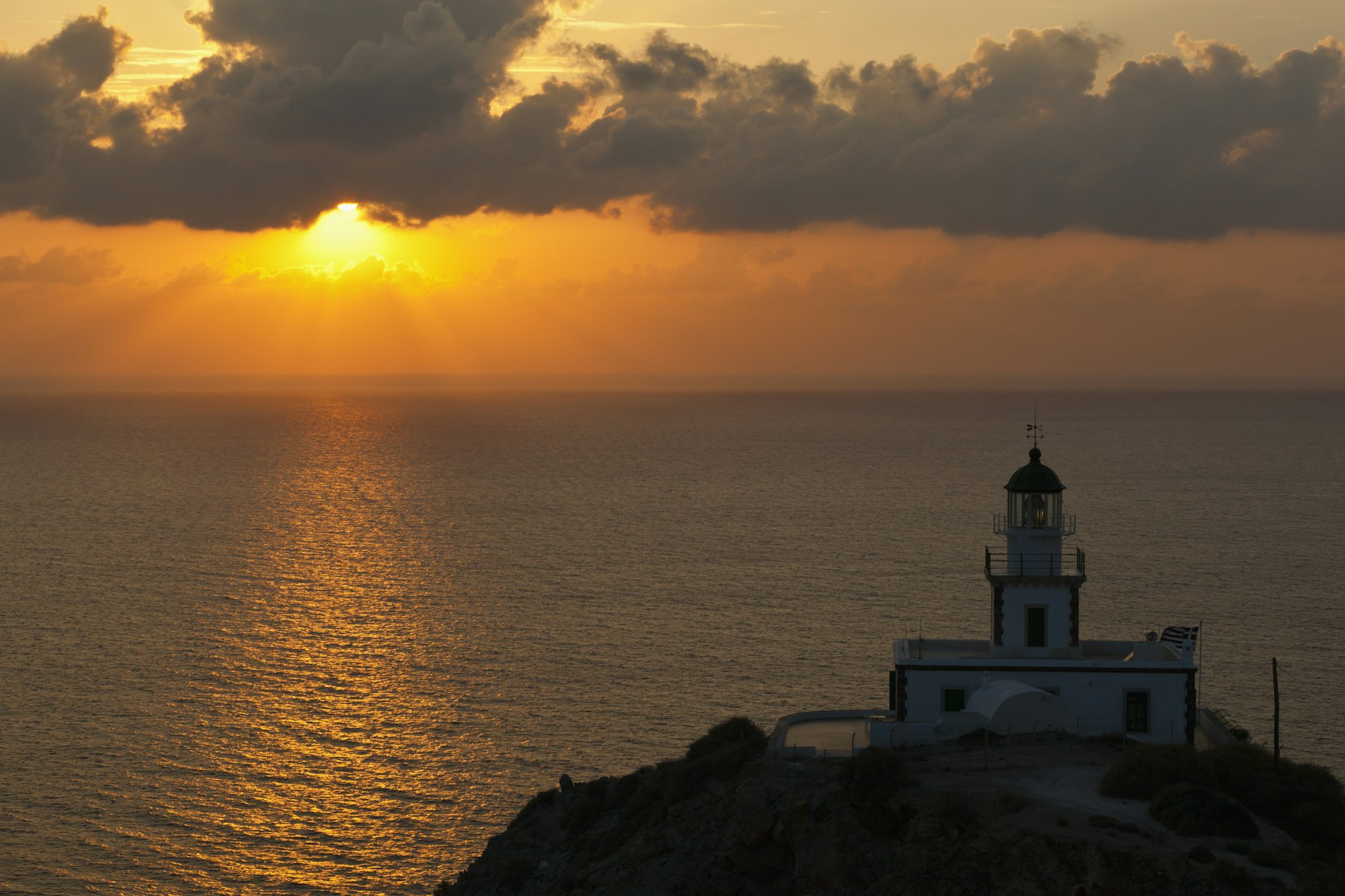 A squat white lighthouse at the edge of a cliff with the sea beyond, and the sky glowing orange as the sun sets