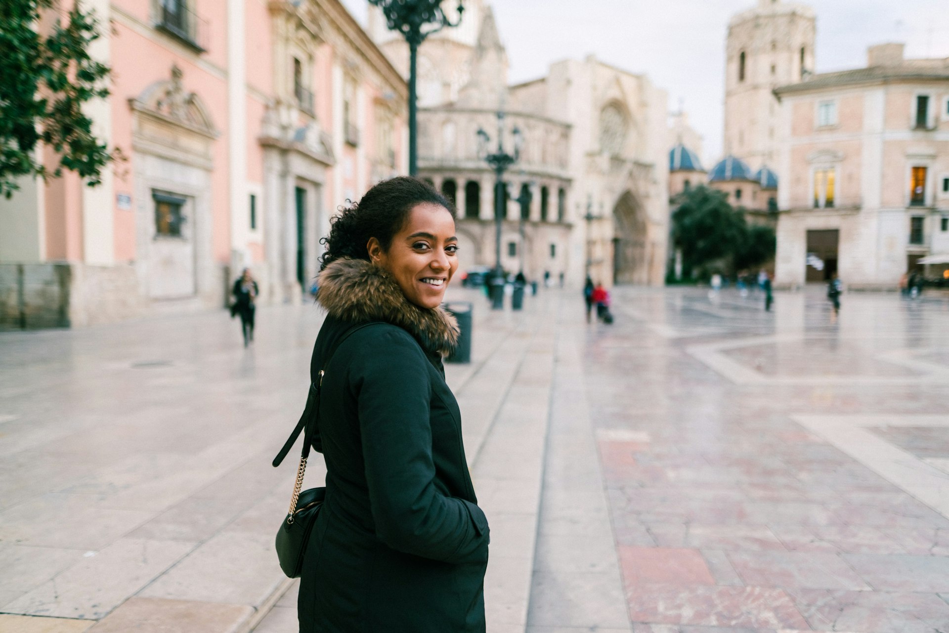 A tourist walking across the Plaza de la Virgen in Valencia turns and faces the camera and smiles