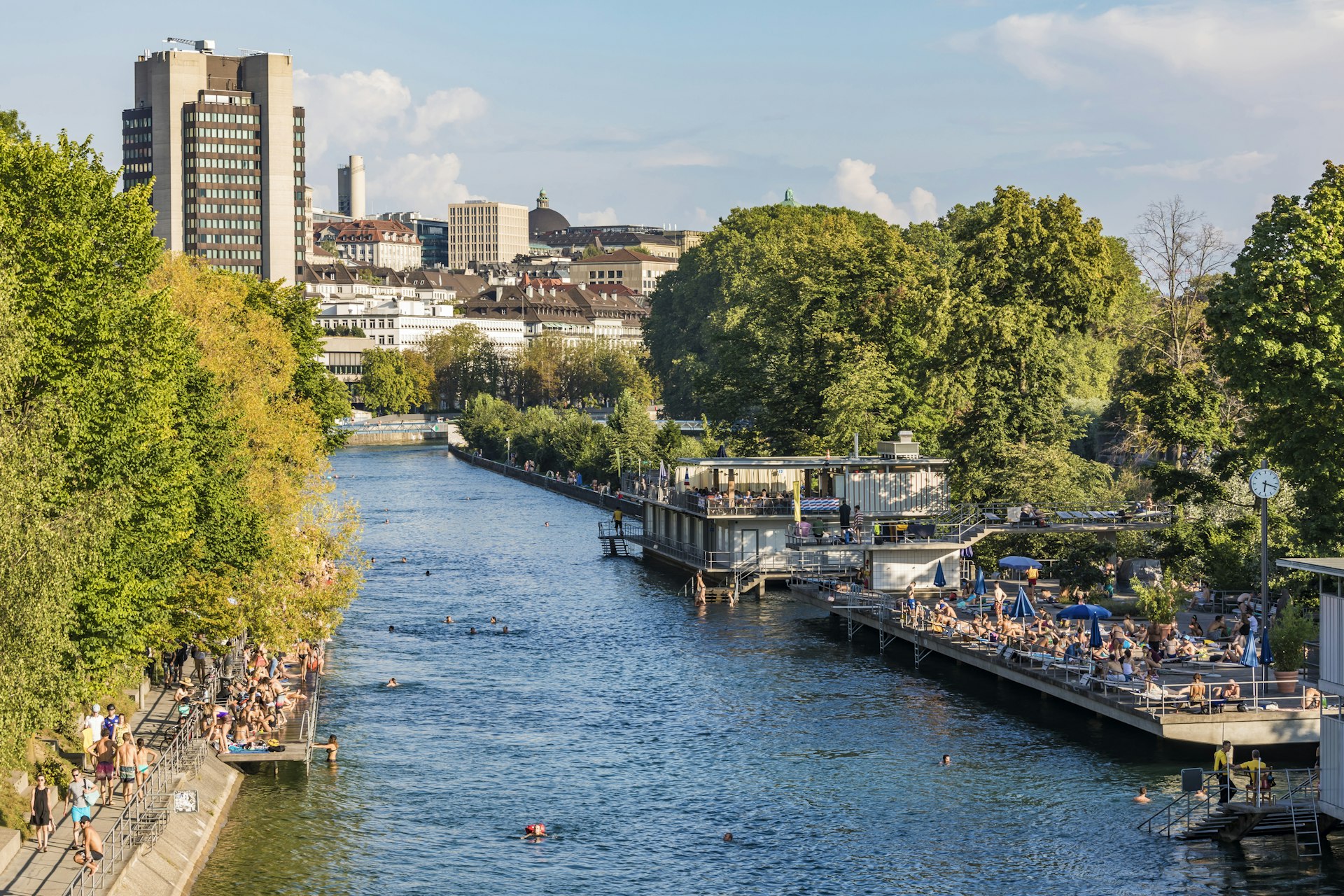 A river running through an urban area. Platforms either side of the river have many people lounging on them, with several swimmers in the river