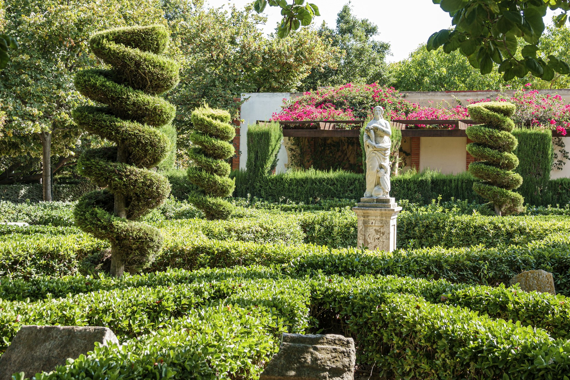 A statue of a woman holding roses in the Viveros Royal Garden, Valencia with three twisting topiary shrubs positioned around the statue and some hanging pink roses in the background. 