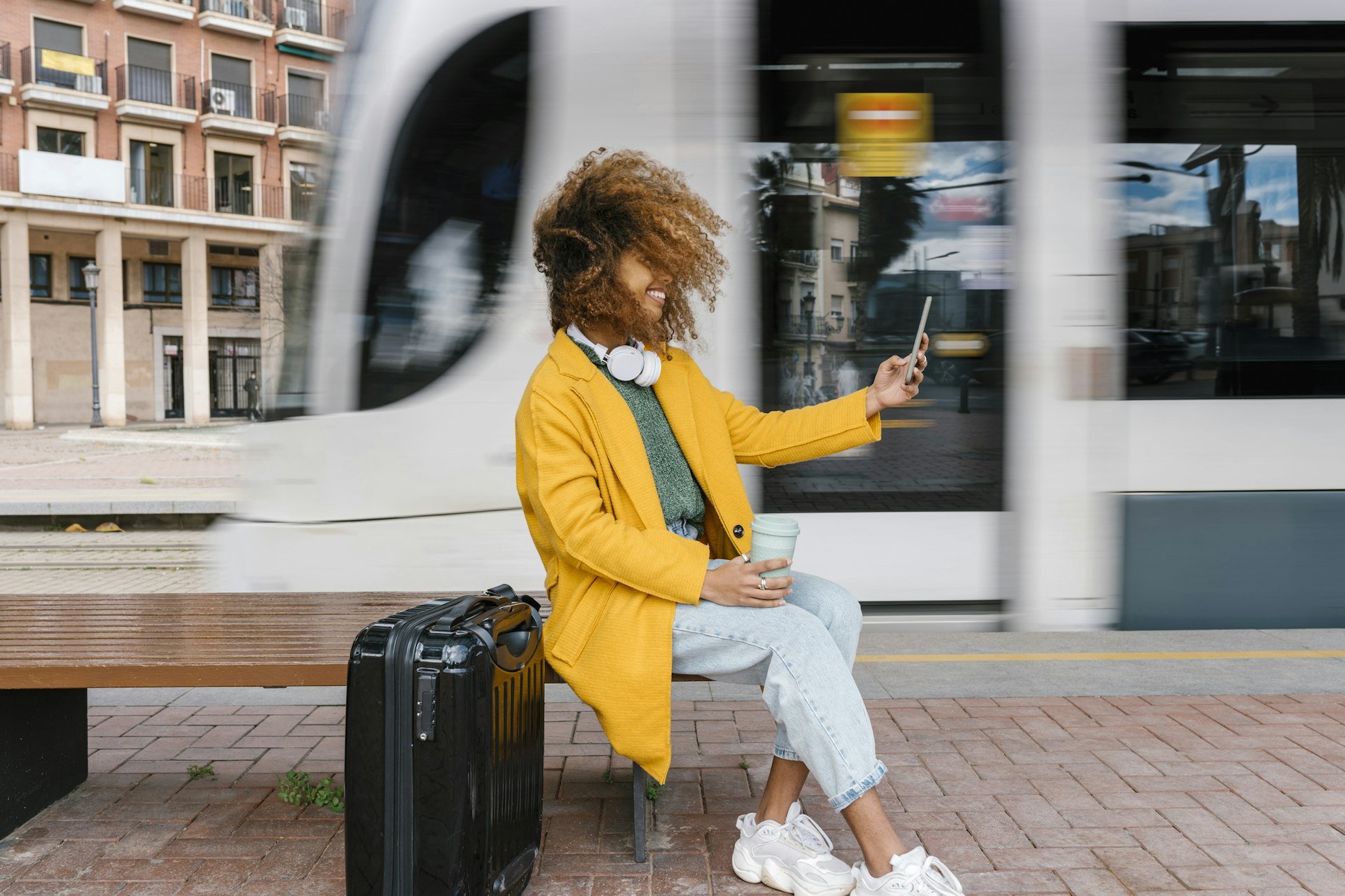 Smiling Afro woman with tousled hair taking selfie through mobile phone while sitting on bench at tram station in Valencia