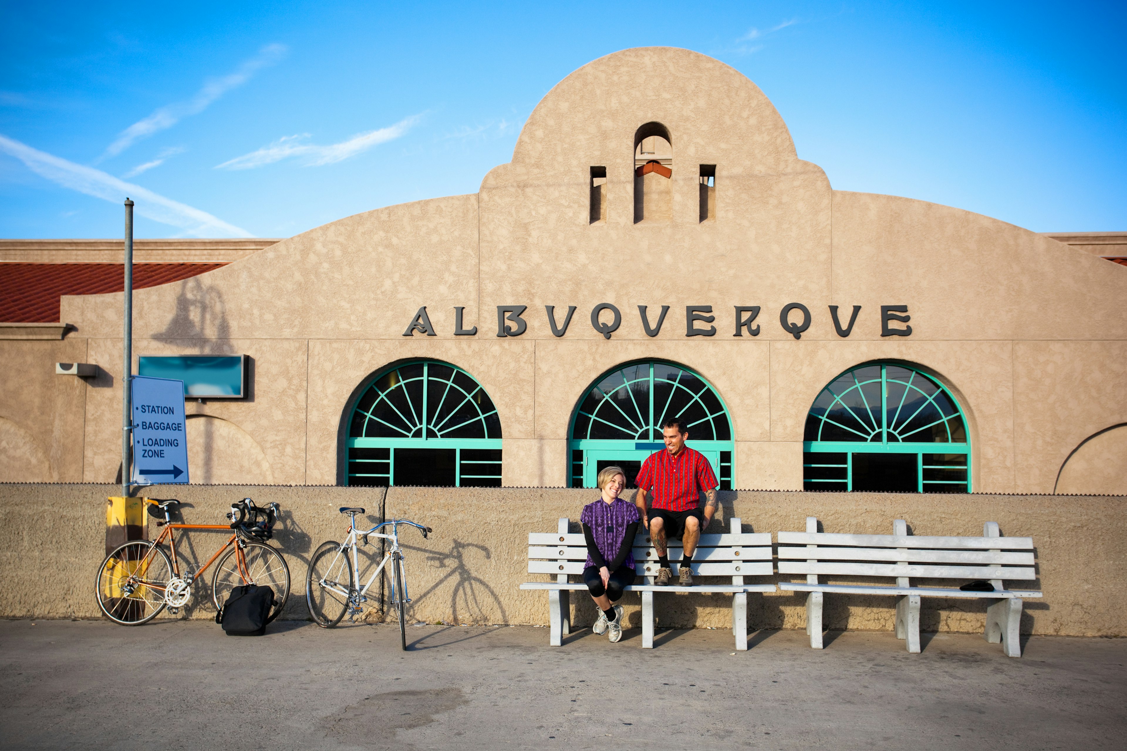 married cyclist couple man and woman take a break and share a laugh while sitting on benches in front of a spanish colonial architecture building train depot.  their bicycles lean against a wall.  horizontal composition taken in albuquerque, new mexico.