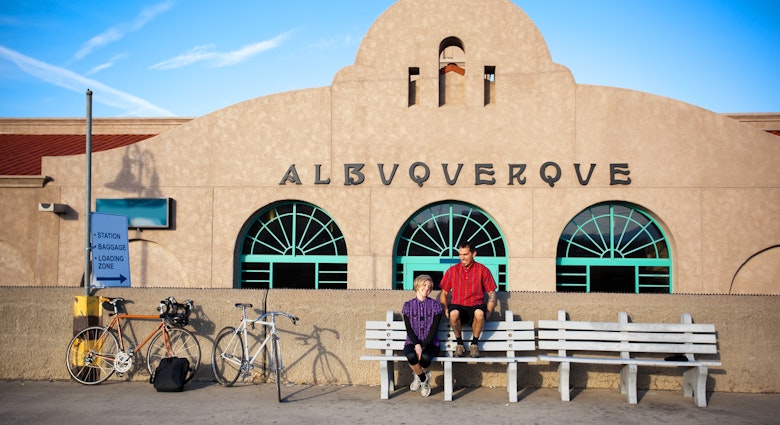 married cyclist couple man and woman take a break and share a laugh while sitting on benches in front of a spanish colonial architecture building train depot.  their bicycles lean against a wall.  horizontal composition taken in albuquerque, new mexico.