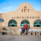married cyclist couple man and woman take a break and share a laugh while sitting on benches in front of a spanish colonial architecture building train depot.  their bicycles lean against a wall.  horizontal composition taken in albuquerque, new mexico.