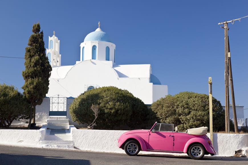 A pink Volkswagen Beetle in front of a chapel on Santorini, Greece