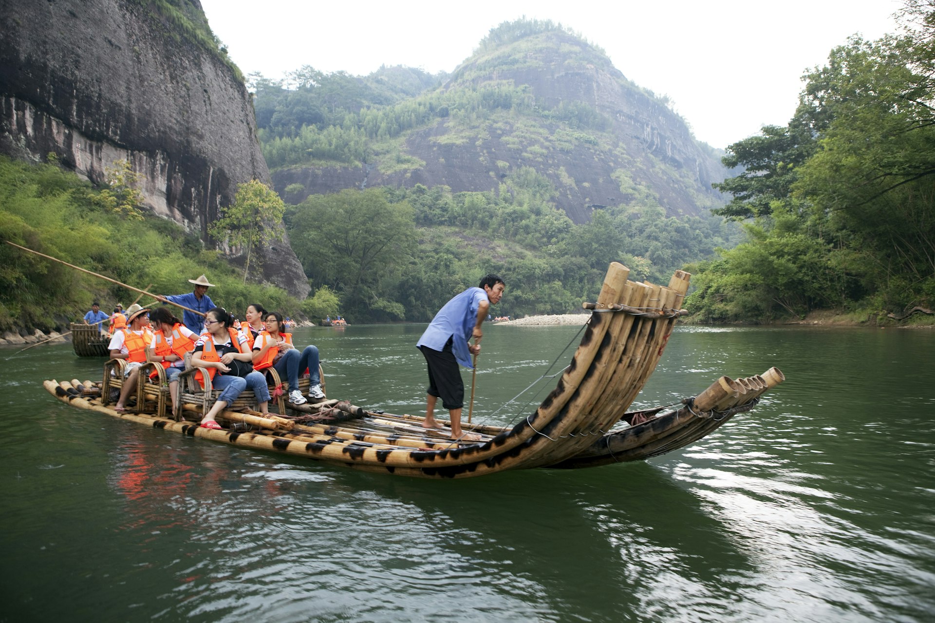 Two bamboo rafts, propelled by men in blue shirts, take a gaggle of visitors in orange life vests, along the Jiuqiu (Nine Bends) River in Wuyi Mountain National Park with tall mountains in the background