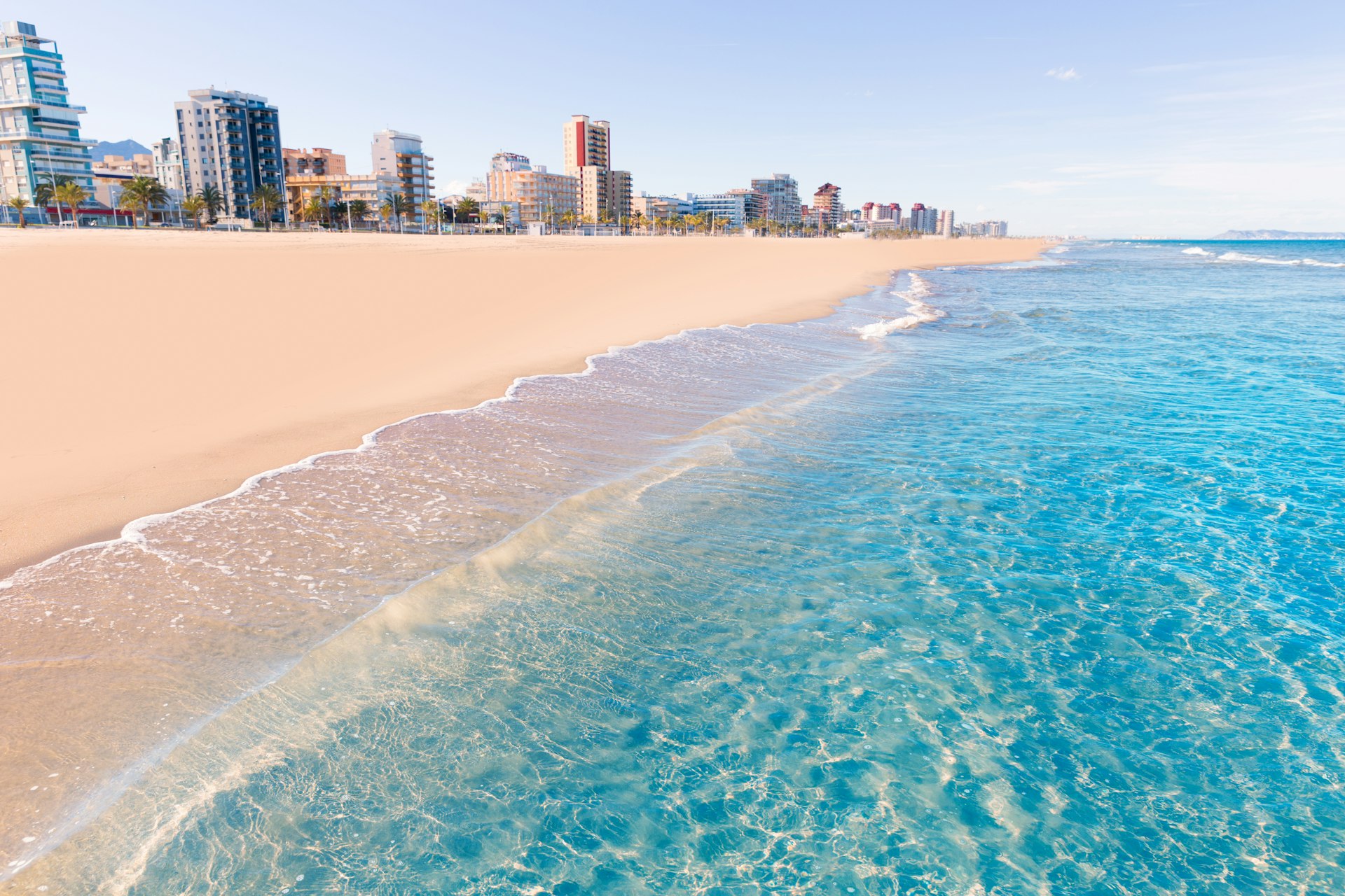 The clear, blue wash of the Mediterranean Sea cuts diagonally against the soft, pinkish sands of Playa de Gandia in Valencia, Spain with several tower blocks rising up in the background