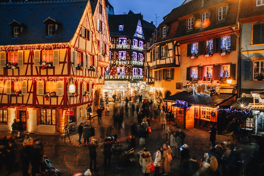 An old town square lined with medieval buildings all lit up with Christmas lights. People are a blur of bodies moving through the Christmas market