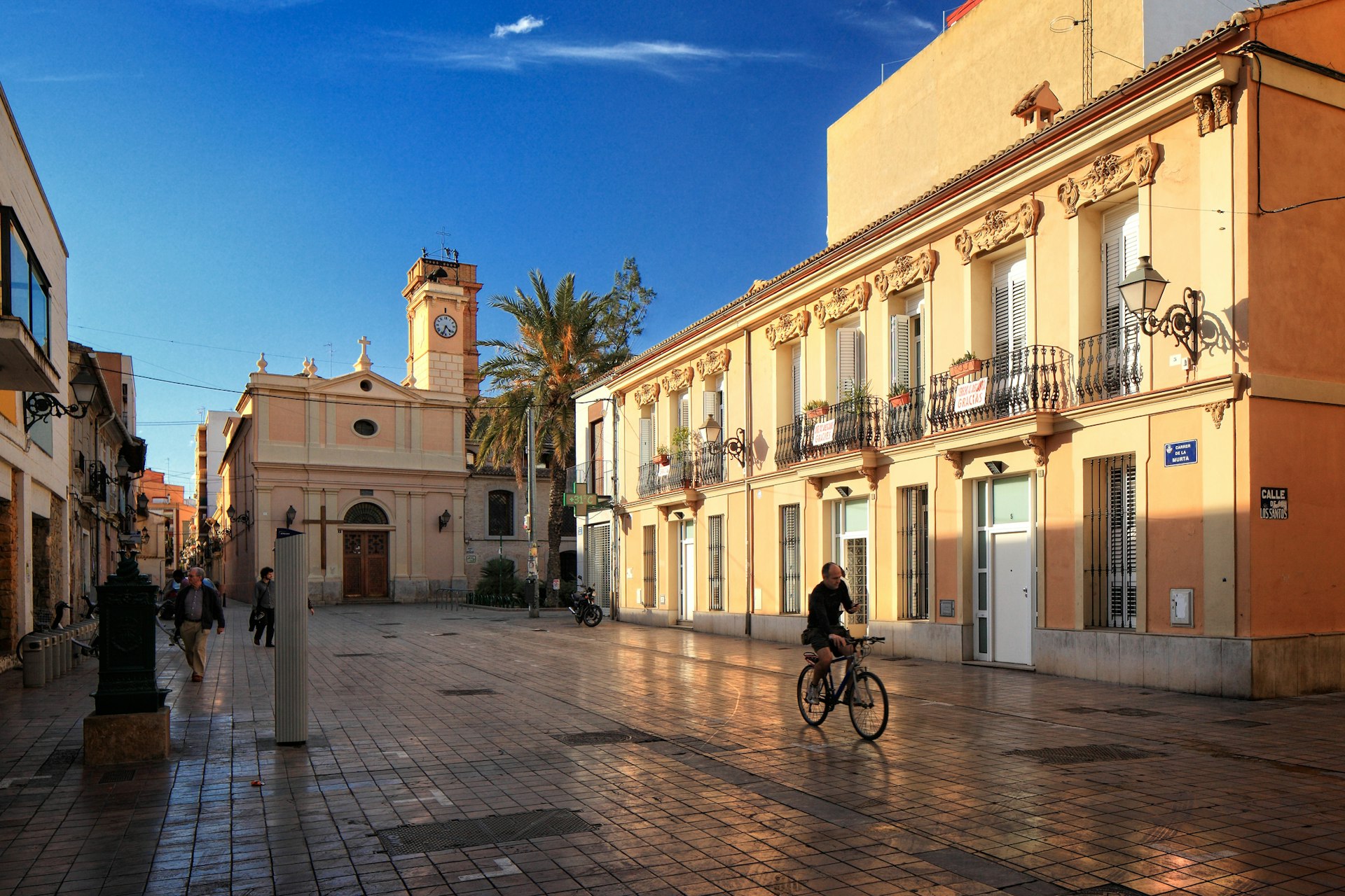 A cyclist whizzes along the shiny tiles of the Benimaclet neighborhood in Valencia, Spain as the sun begins to lift over the buildings on the left to illuminate the lovely balconies on a wonderful peach-coloured building.
