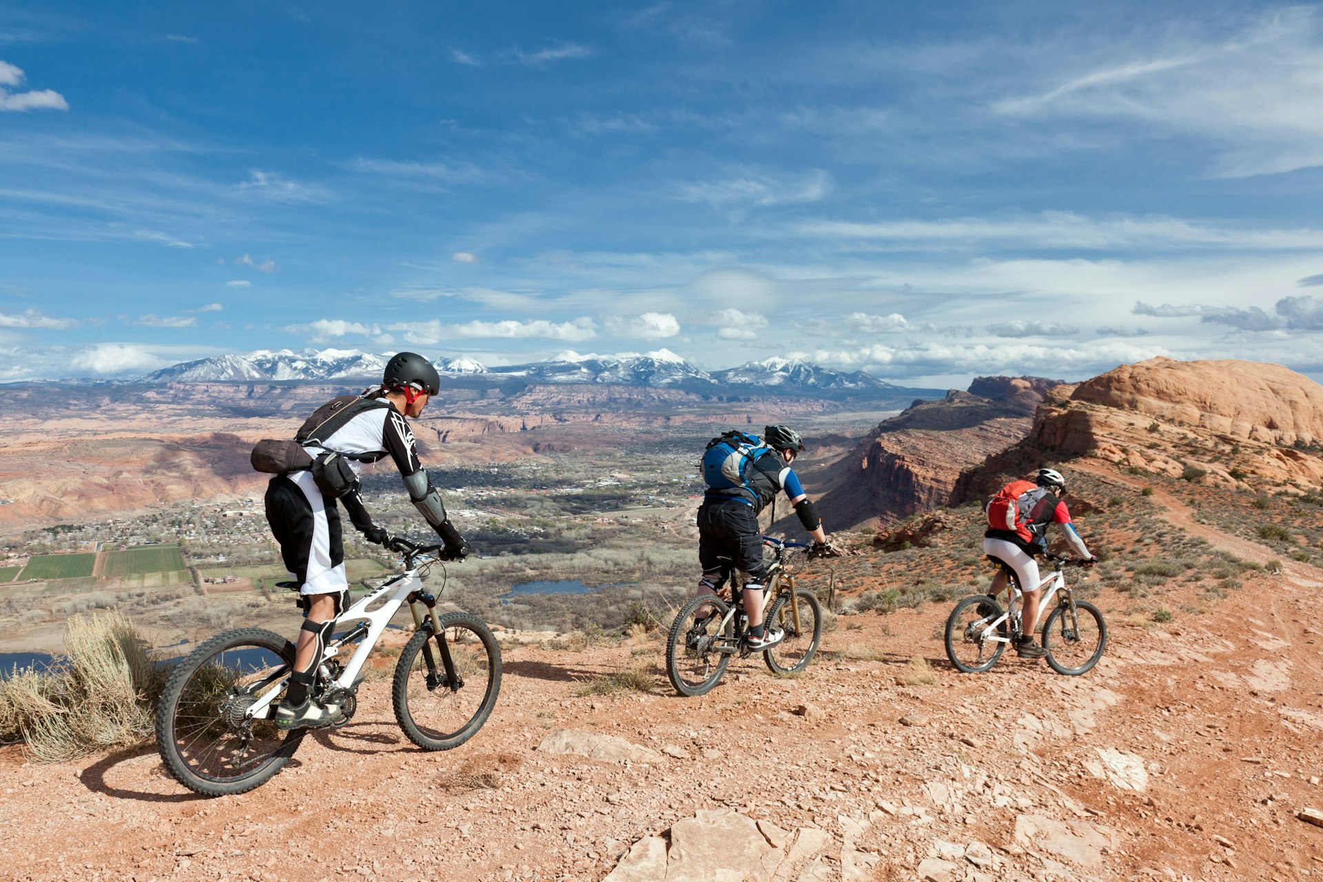 Three people bike on a ridge in Moab, with red rock formations visible in the distance