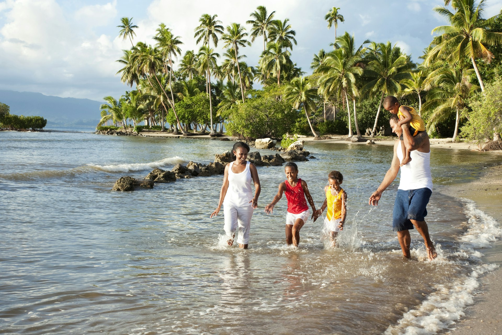 A smiling Fijian family of three kids and a mum and dad playing in the sea on a white-sand beach with palm trees behind them on Fiji's Vanua Levu