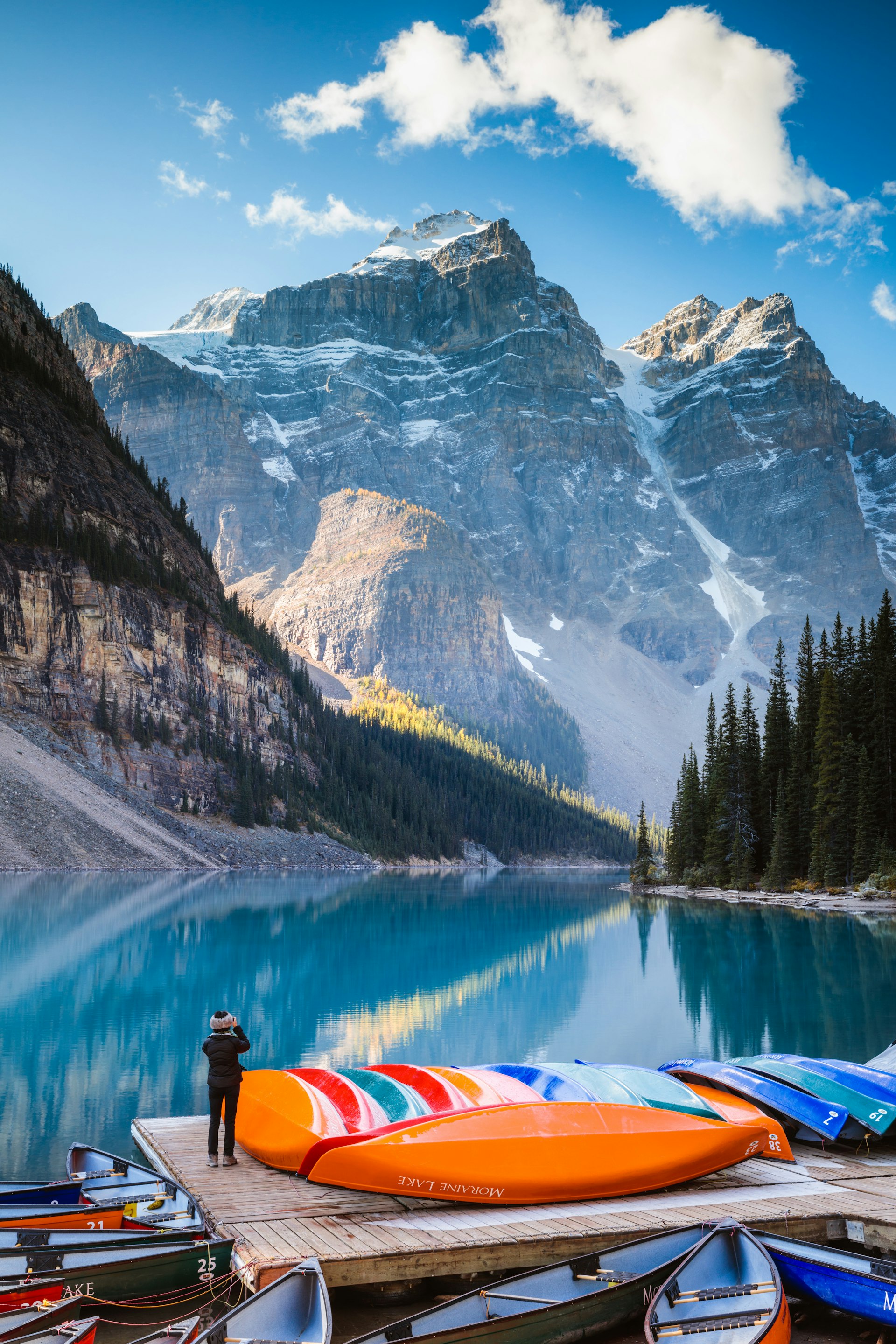 Woman taking a photo of Moraine Lake, in Banff National Park, next to a stack of colorful canoes in autumn. 