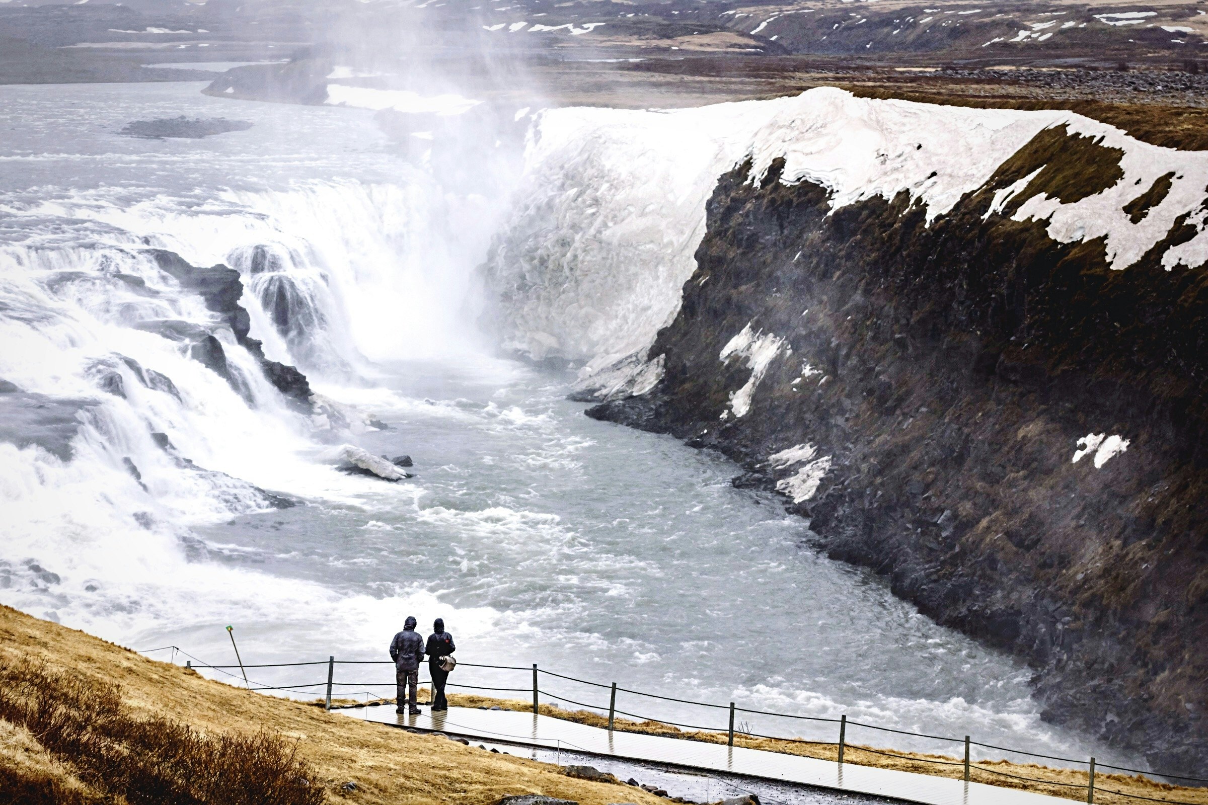 Two people stand overlooking Gullfloss waterfall in Iceland. The falls are huge, with vast amounts of water pouring from them into the lake below.