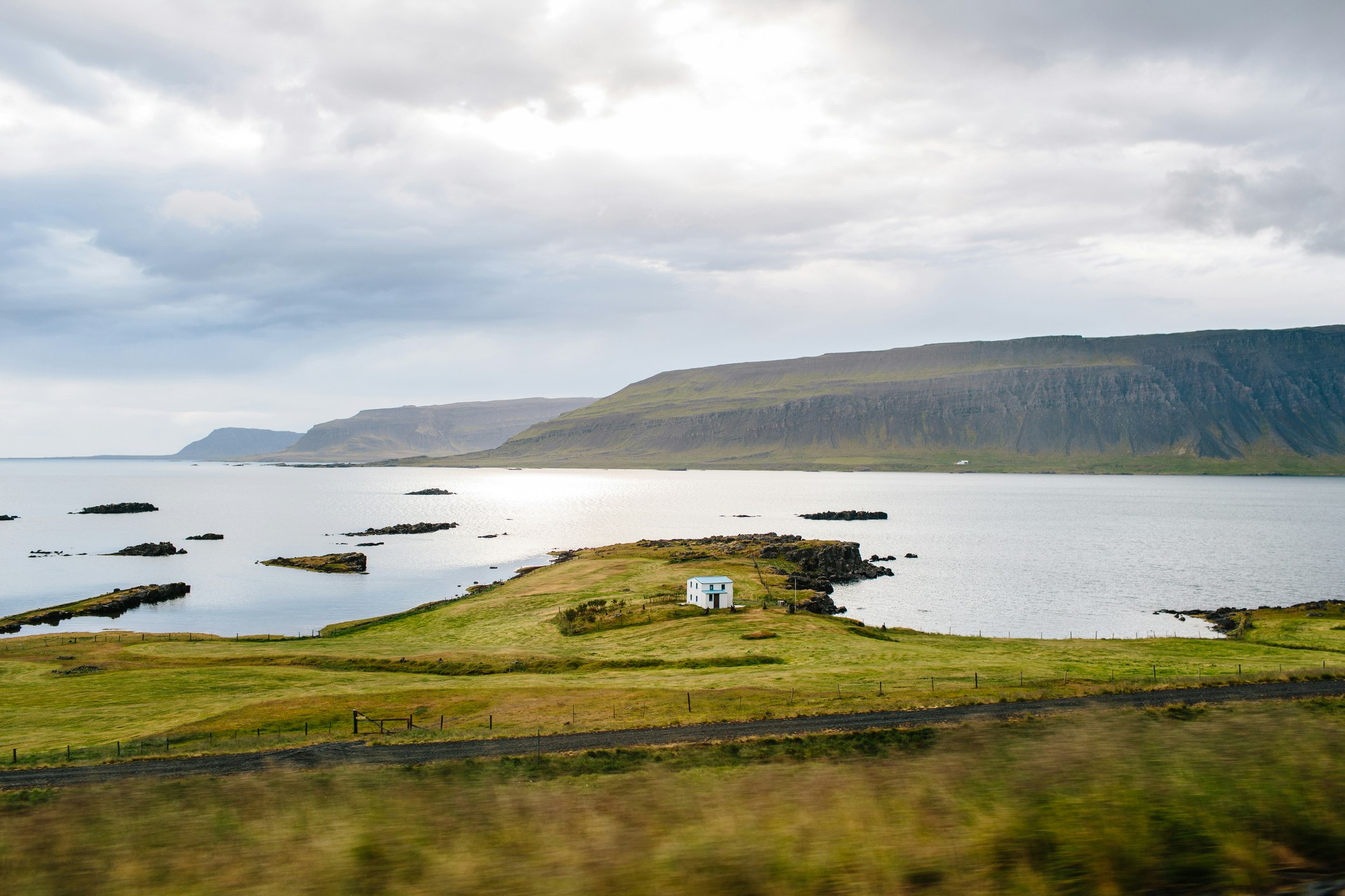 A lone white house stands on a small grassy hillside in the Westfjords, Iceland. Behind the house is the blue sea.