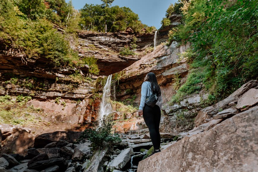 A woman is standing on a rocky step looking upwards at Kaaterskill Falls