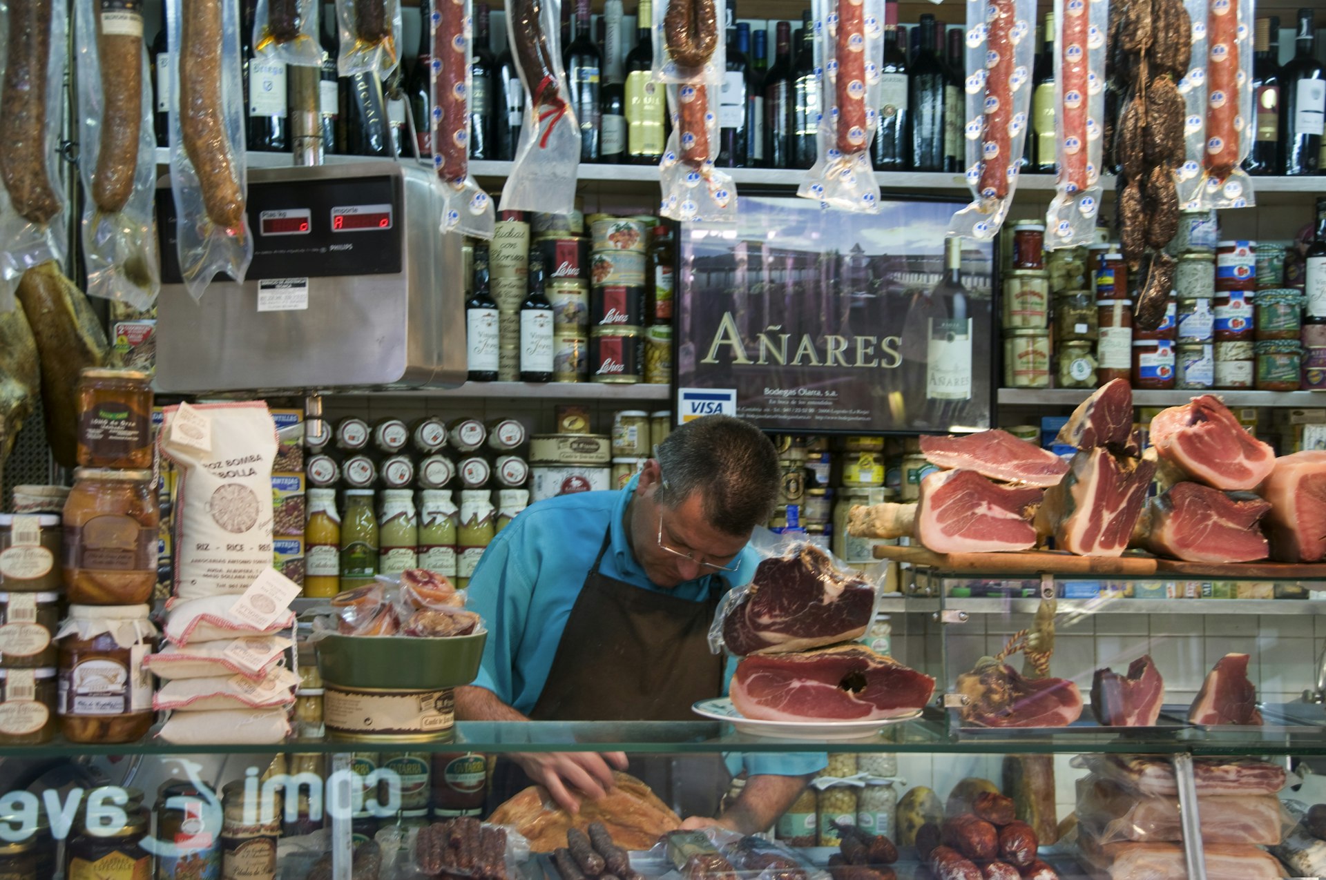 A man in a market stall with jamón on the counter and sausages dangling overhead