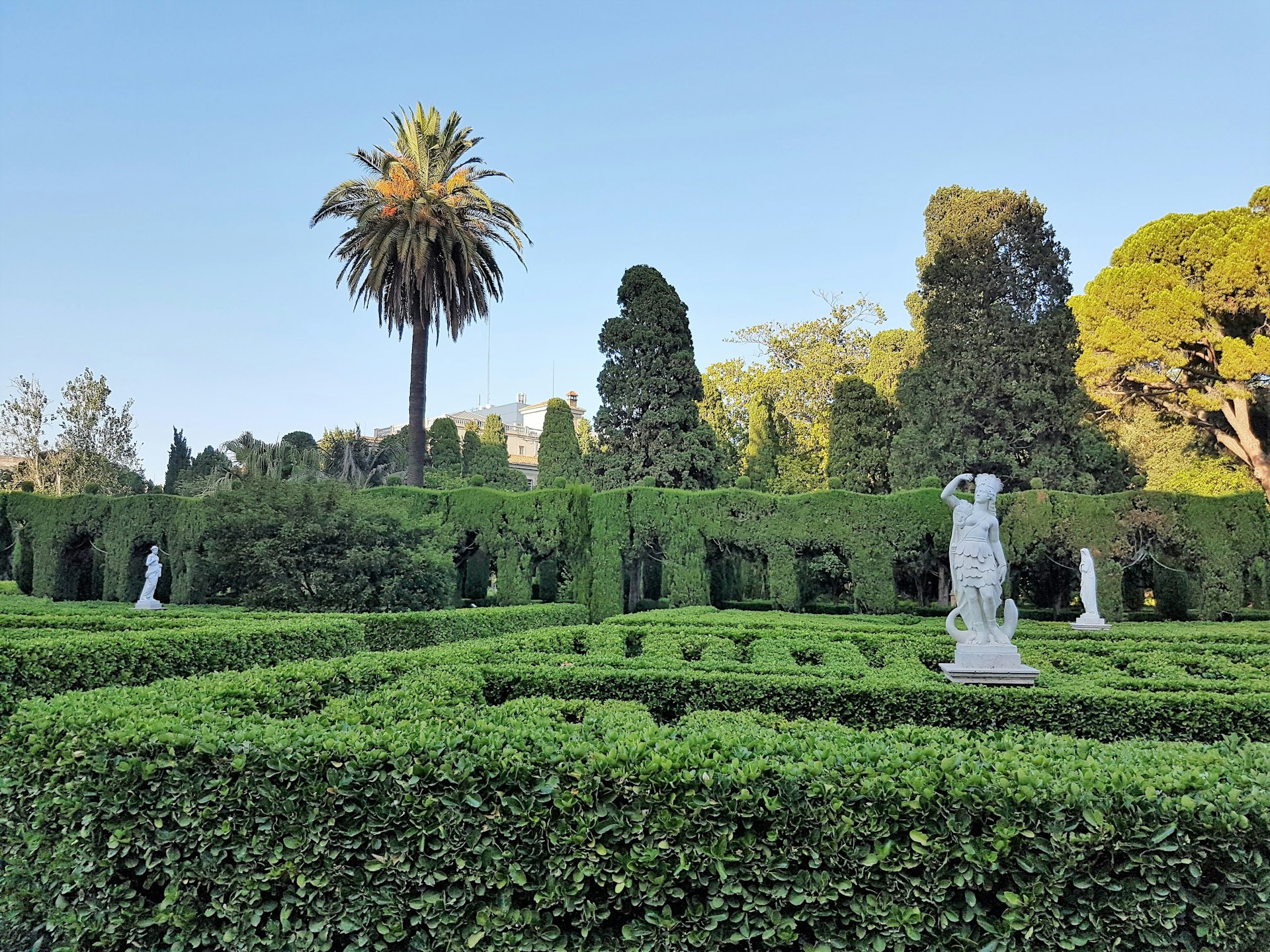 A neoclassical garden featuring manicured hedges and a marble sculpture with palm trees in the background