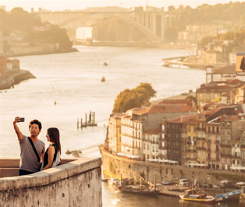 A couple takes a selfie at the Jardim do Morro. In the background, a view of the cityscape of Porto is visible, including its famous bridges.