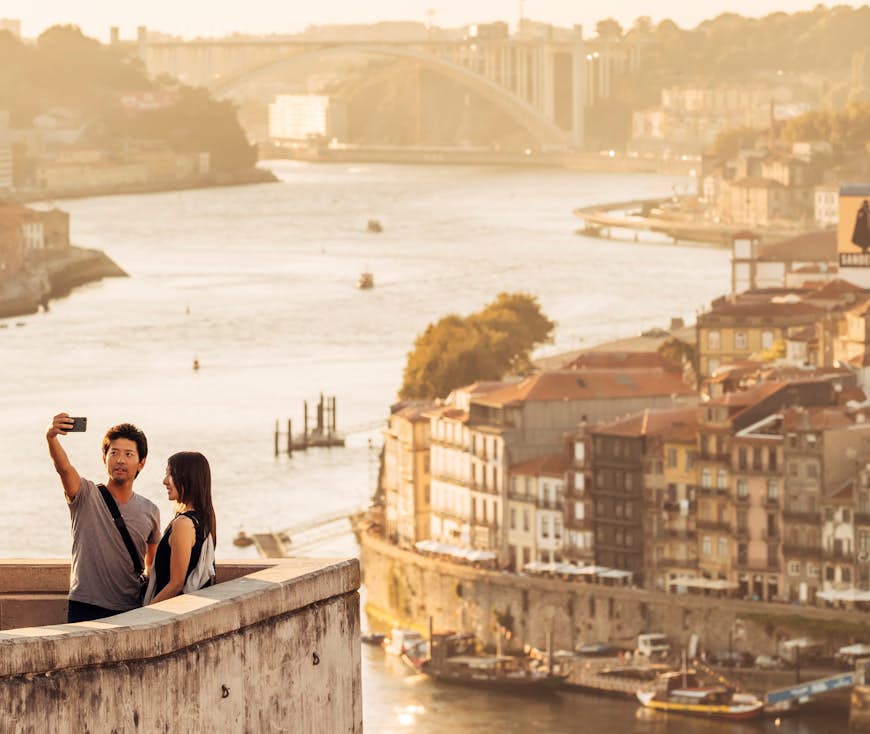 Ett par tar en selfie på Jardim do Morro.  I bakgrunden syns en utsikt över stadsbilden i Porto, inklusive dess berömda broar.
