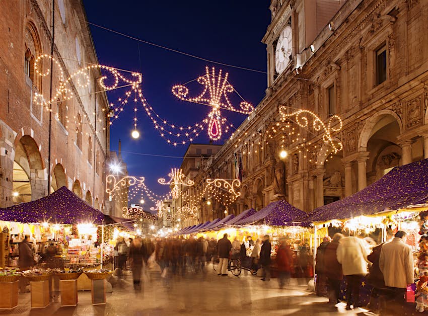 People walk the streets and browse stalls in one of Milan's Christmas markets.  The stalls are illuminated by bright fairy lights.