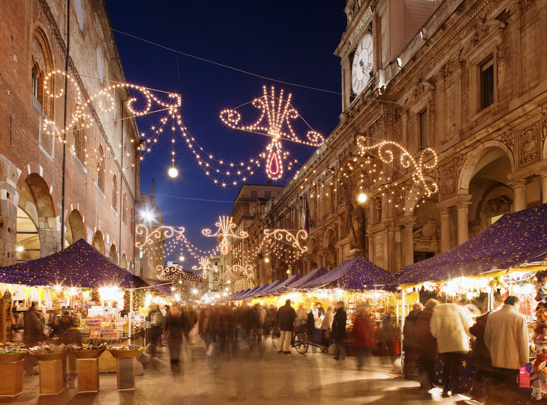People walk the streets and browse stalls in one of Milan's Christmas markets. The stalls are illuminated by bright fairy lights.