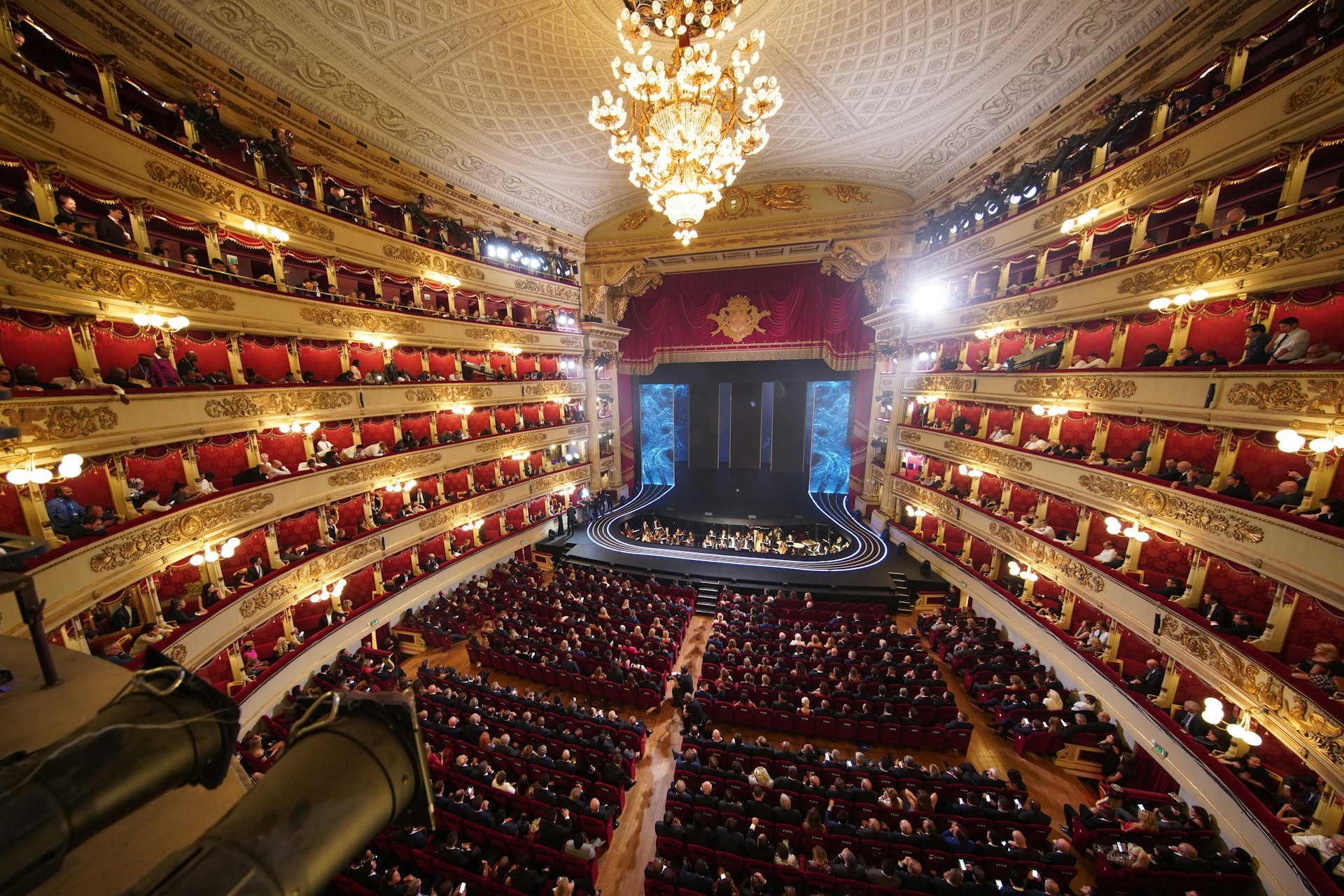 A view from the stands of the large Teatro alla Scala opera house in Milan. The venue has lots of seating and a lit stage in the centre.