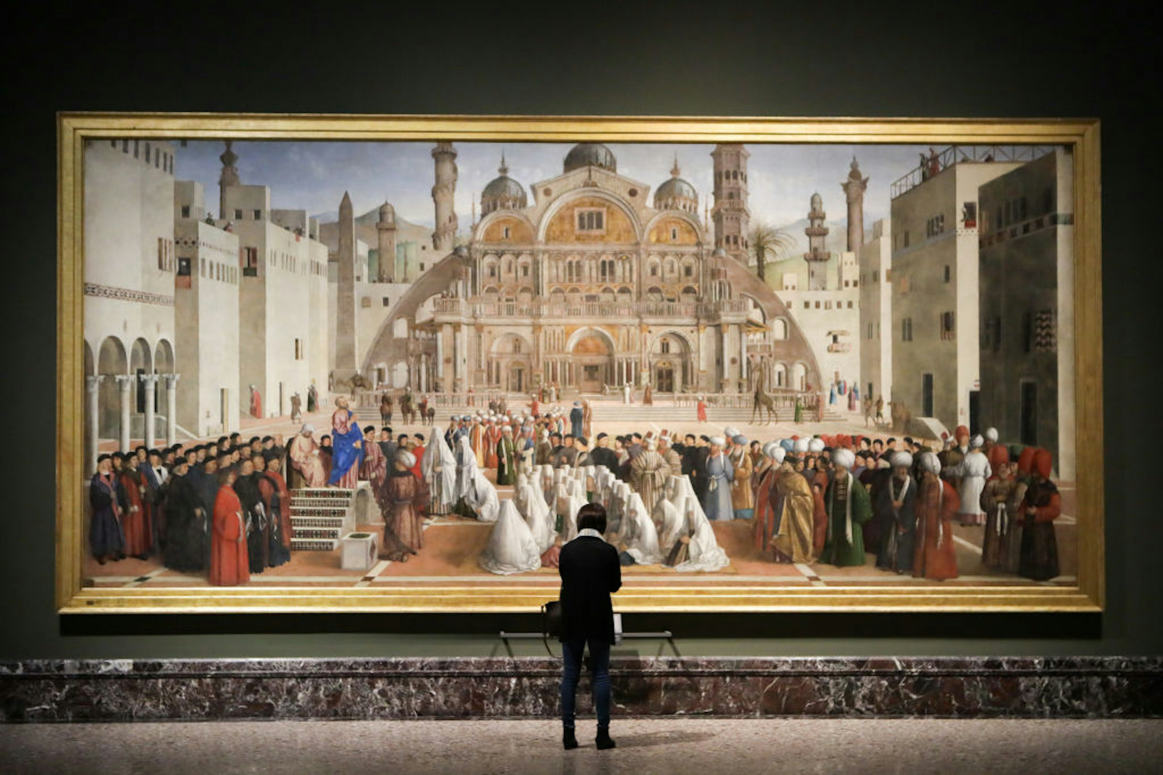 A lady stands in front of a huge painting, which hands on the wall of the Pinacoteca di Brera in Milan.