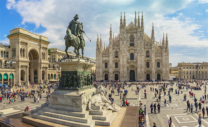 A view of the Piazza del Duomo, the Cathedral and equestrian monument to Vittorio Emanuele II in Milan.  Crowds of people walk around the large square.