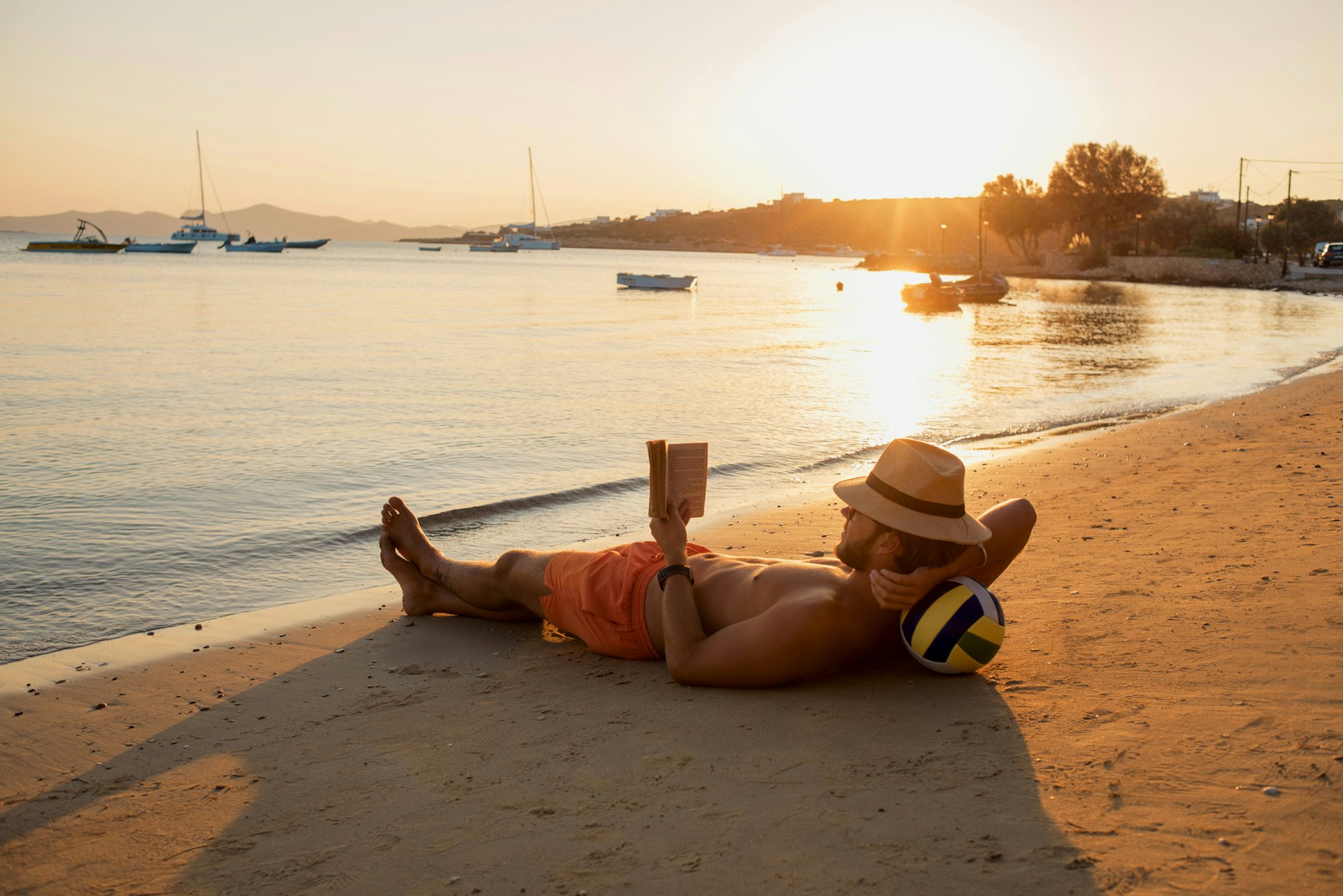 A man is lying on a sandy beach reading a book as the sun sets in Paros