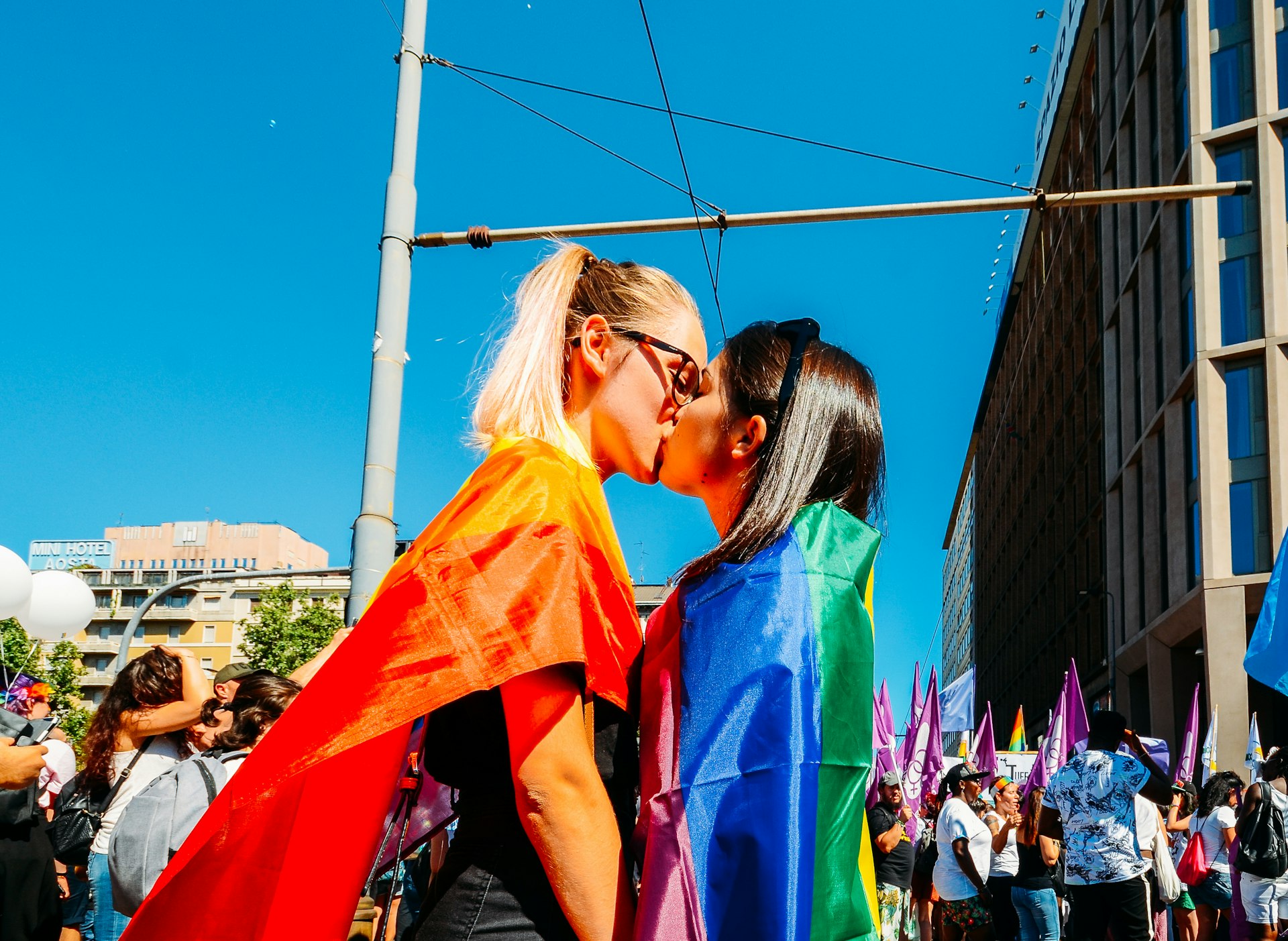 Women kissing at the Milan Pride March with rainbow flags draped over their shoulders