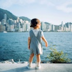 Rear view of cute little Asian girl with sunglasses enjoying the sun by the promenade and looking over city skyline and Victoria harbor on a lovely sunny day Hong Kong.