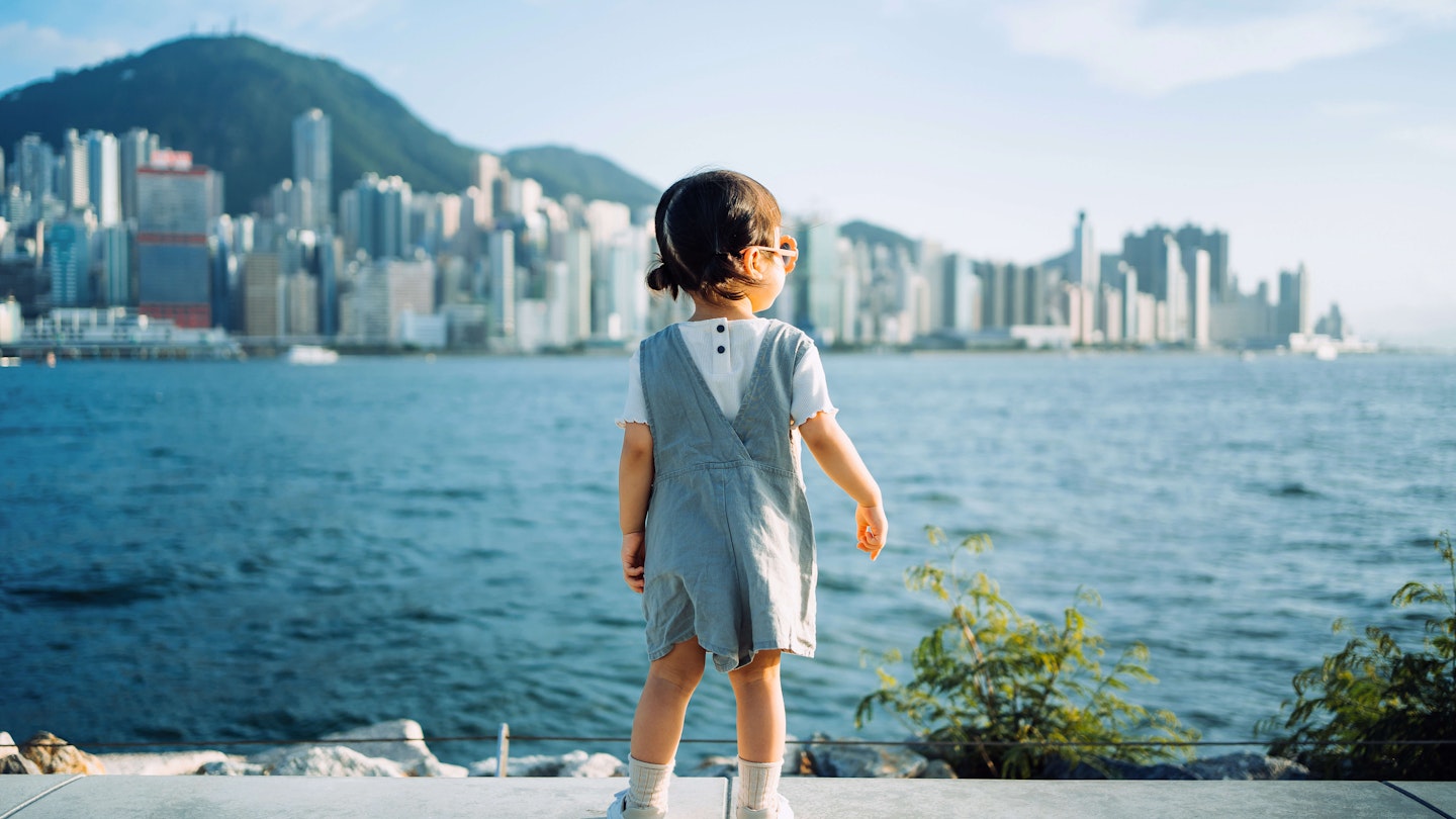 Rear view of cute little Asian girl with sunglasses enjoying the sun by the promenade and looking over city skyline and Victoria harbor on a lovely sunny day Hong Kong.