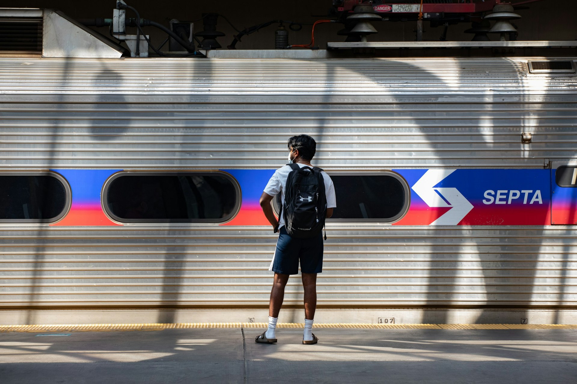 A man wearing shorts and a backpack stands on the platform in front of the SEPTA train car. 