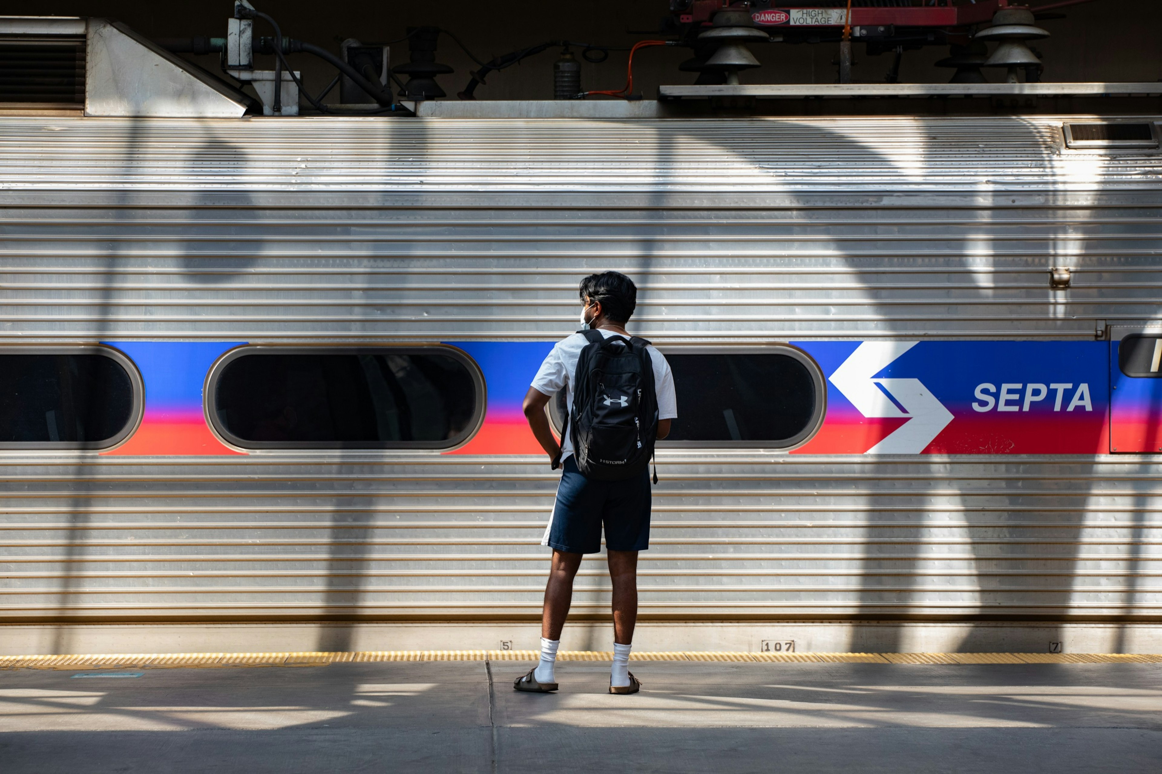 A man wearing shorts and a backpack stands on the platform in front of the SEPTA train car in Philadelphia, Pennsylvania