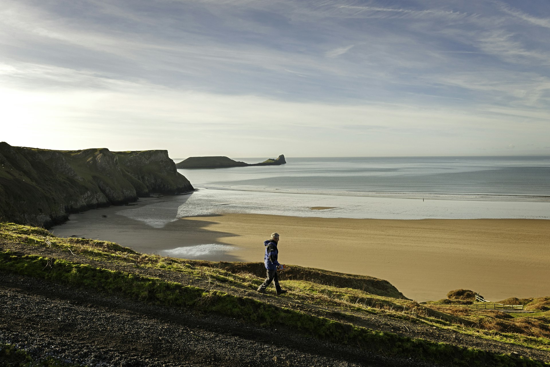 Man walking above Rhossili Beach, with Worm's Head in the background 