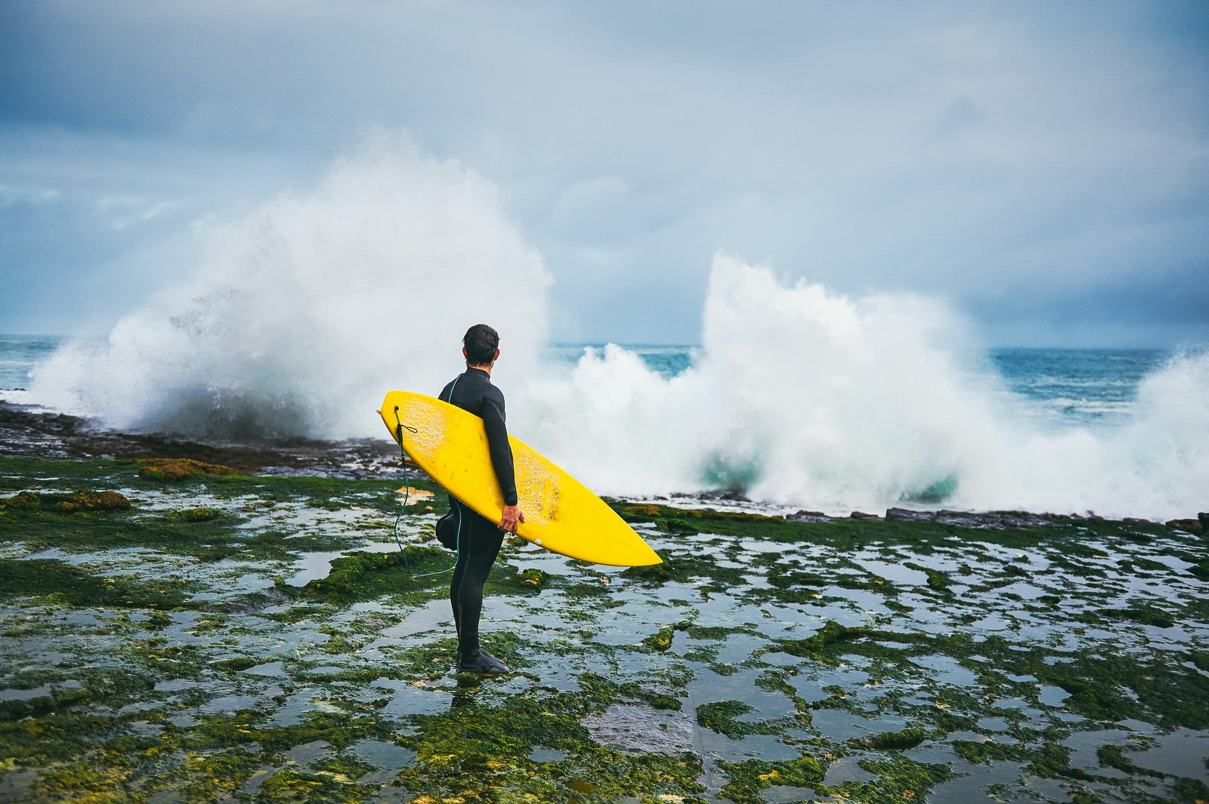 Surfer in a full wet suit holding a yellow surfboard looking out towards huge Atlantic waves hitting the coast of Ireland