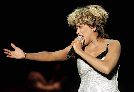 Tina Turner holds a microphone will performing on stage in a silver dress. 