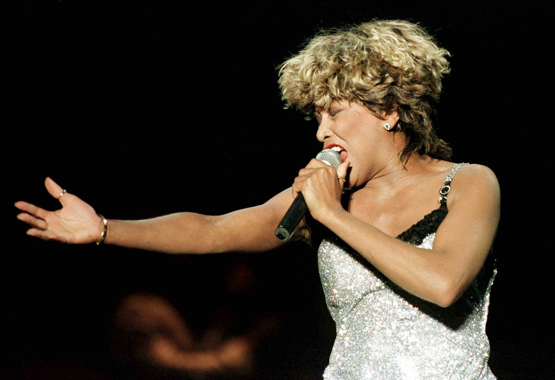 Tina Turner holds a microphone will performing on stage in a silver dress. 