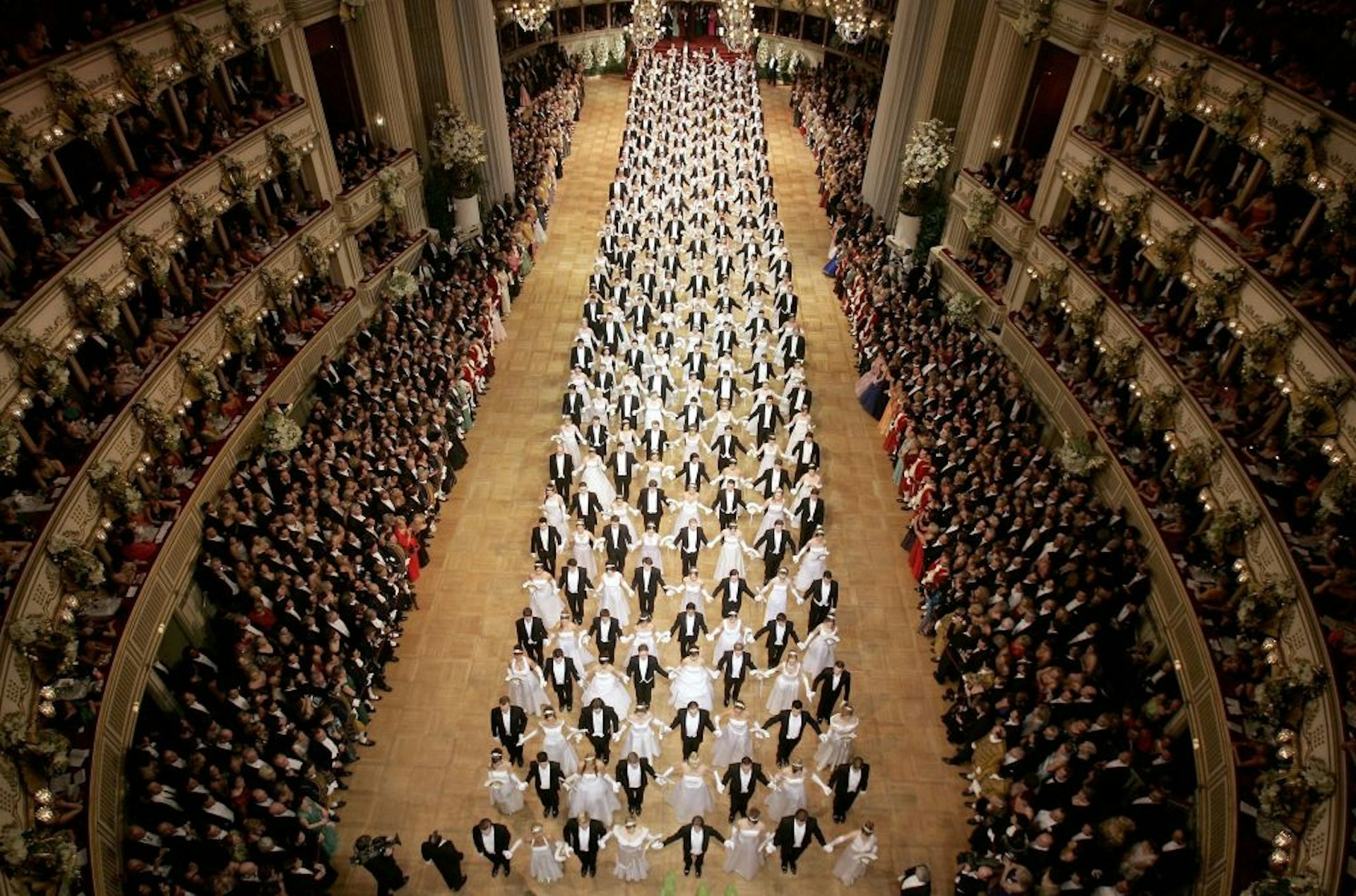 A top-down view of the Vienna Opera Ball, with men, dressed in black, and women, dressed in white, creating a crisscross pattern across the grand venue's large dancefloor.