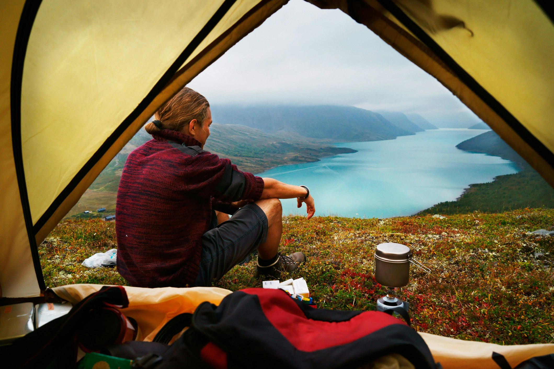 A young hiker sits outside his tent on a grassy spot overlooking Lake Gjende, Jotunheimen National Park, Norway.