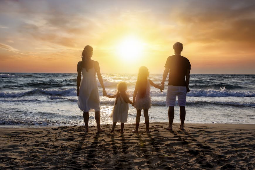 Rear View Of Parents Holding Hands With Daughters Standing At Beach Against Orange Sky
