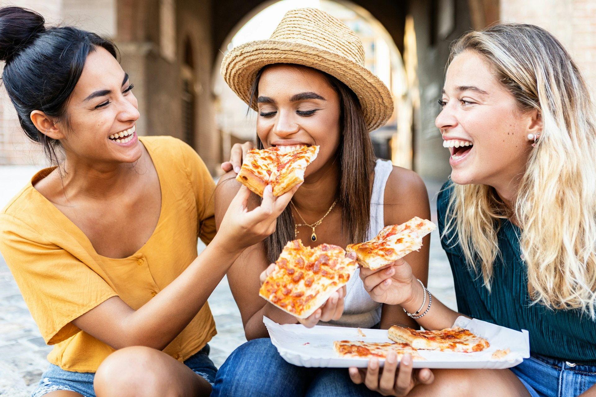Three cheerful multiracial women eating pizza in the street in Florence