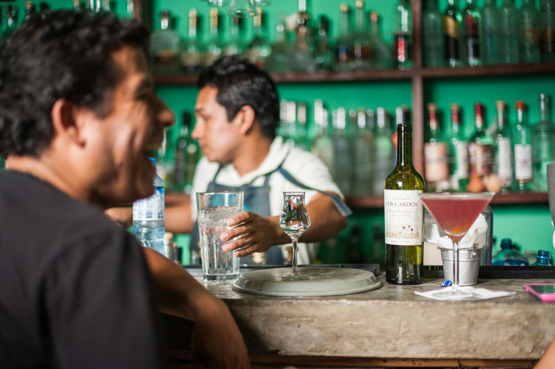 Vintage South American bar, people drinking cocktails, being served by the bartender. Alcoholic beverage bottles and drink glasses displayed in the background