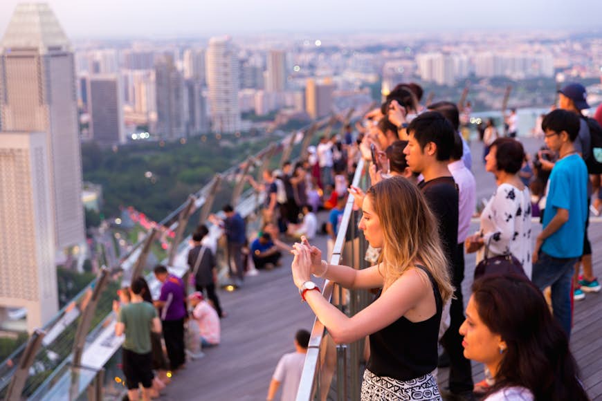 People line a high balcony taking photographs as the sun sets over the city skyline