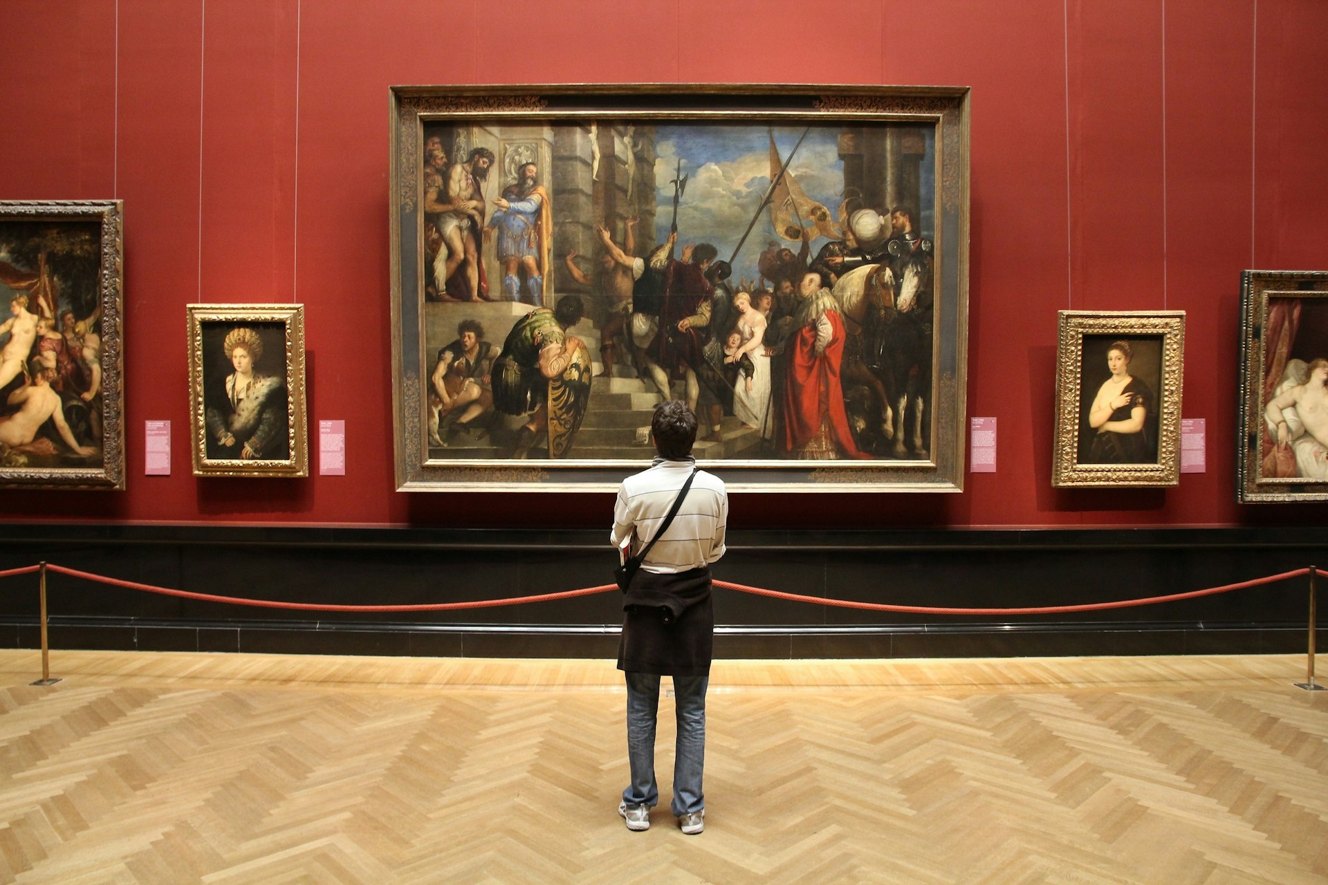 A tourist admires art in the Museum of Art History in Vienna. The male tourist stands with his back to the camera, with a huge painting in a large frame in front of him.