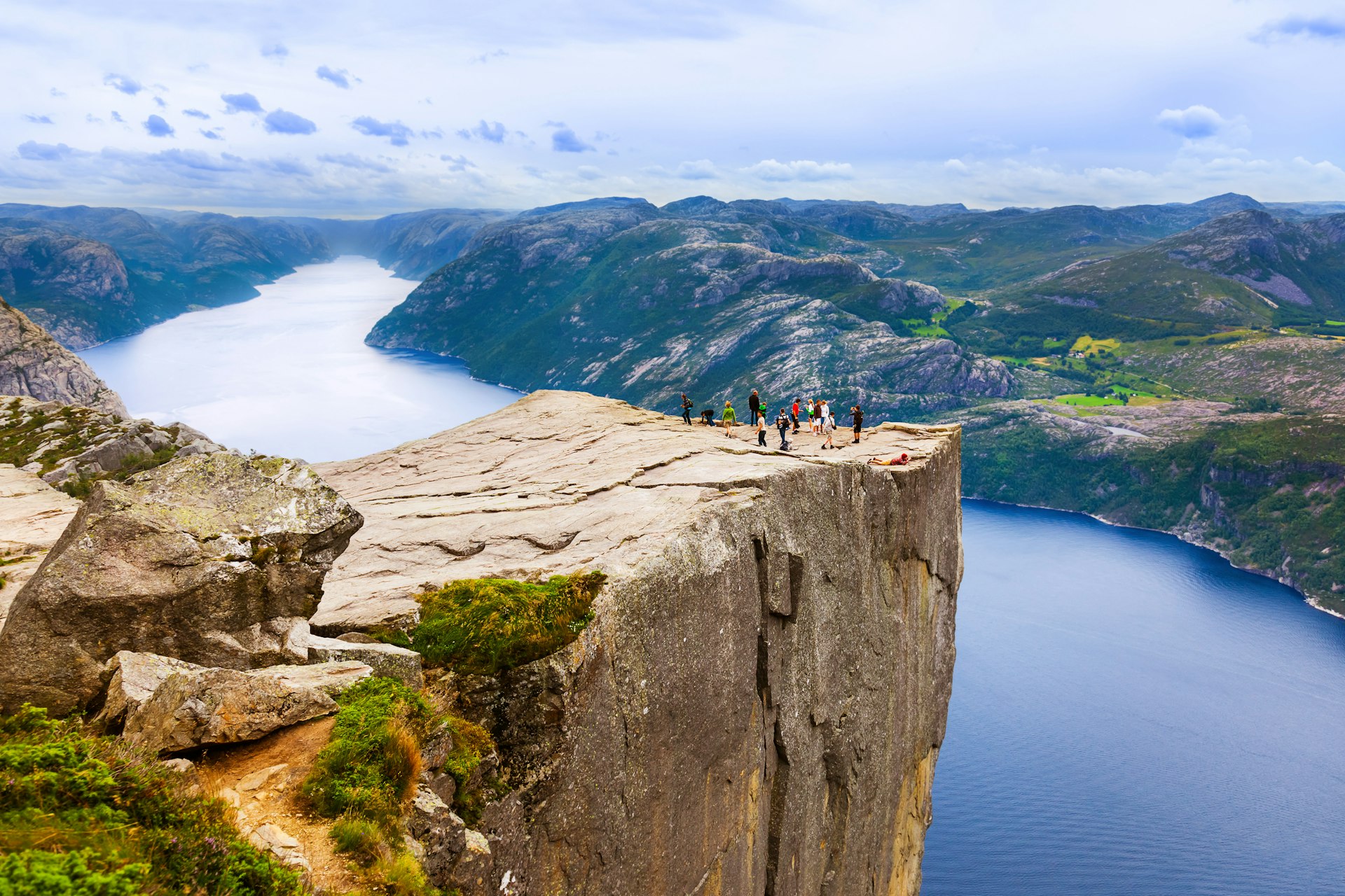 The Preikestolen in fjord Lysefjord: a large rocky platform sticking out from a cliff face in Norway. People stand on the cliff to observe the view of the large lake below.