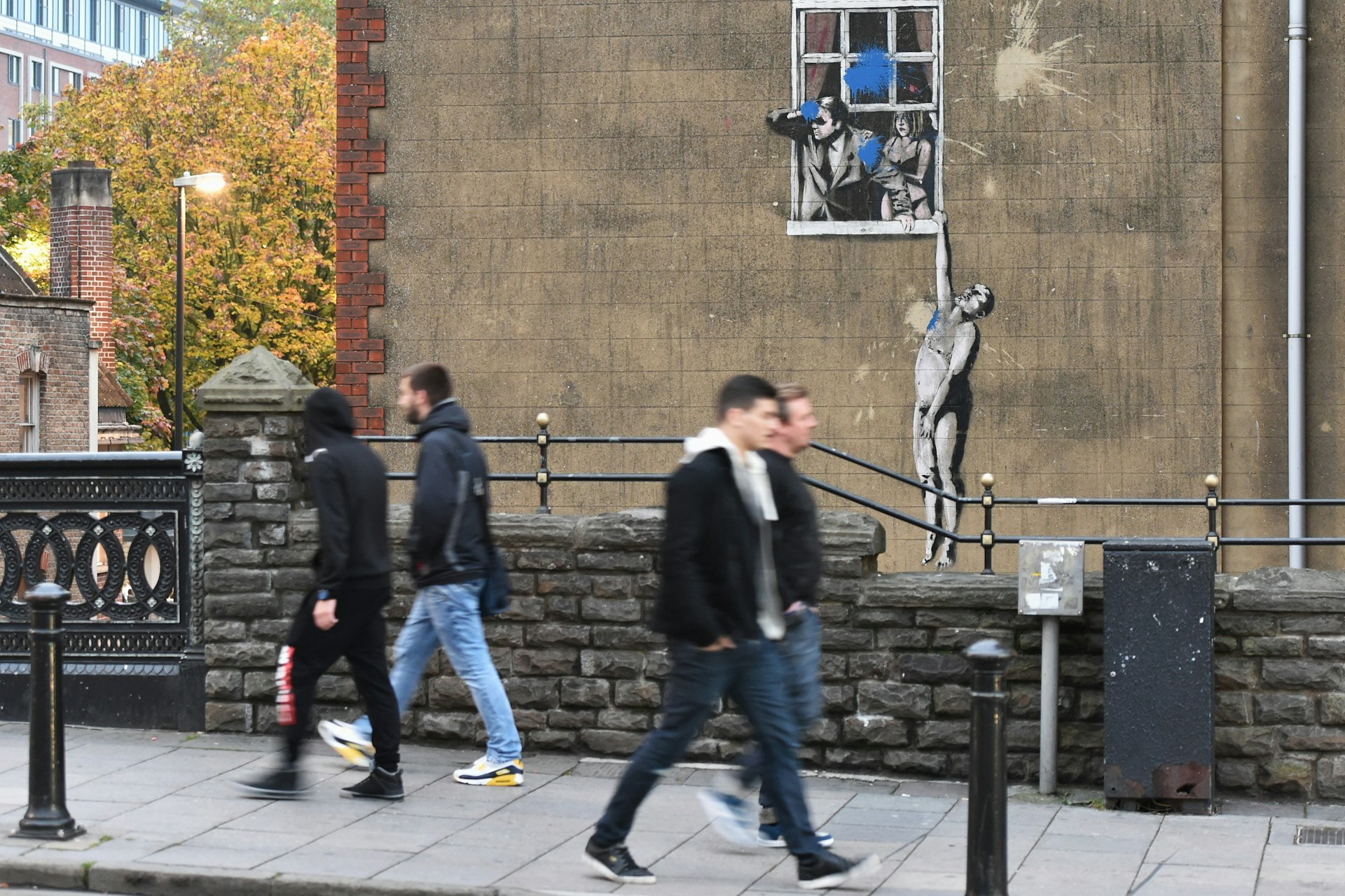Graffiti on a wall of a man looking out of a window with a woman standing behind him. A naked man dangles from the window ledge 