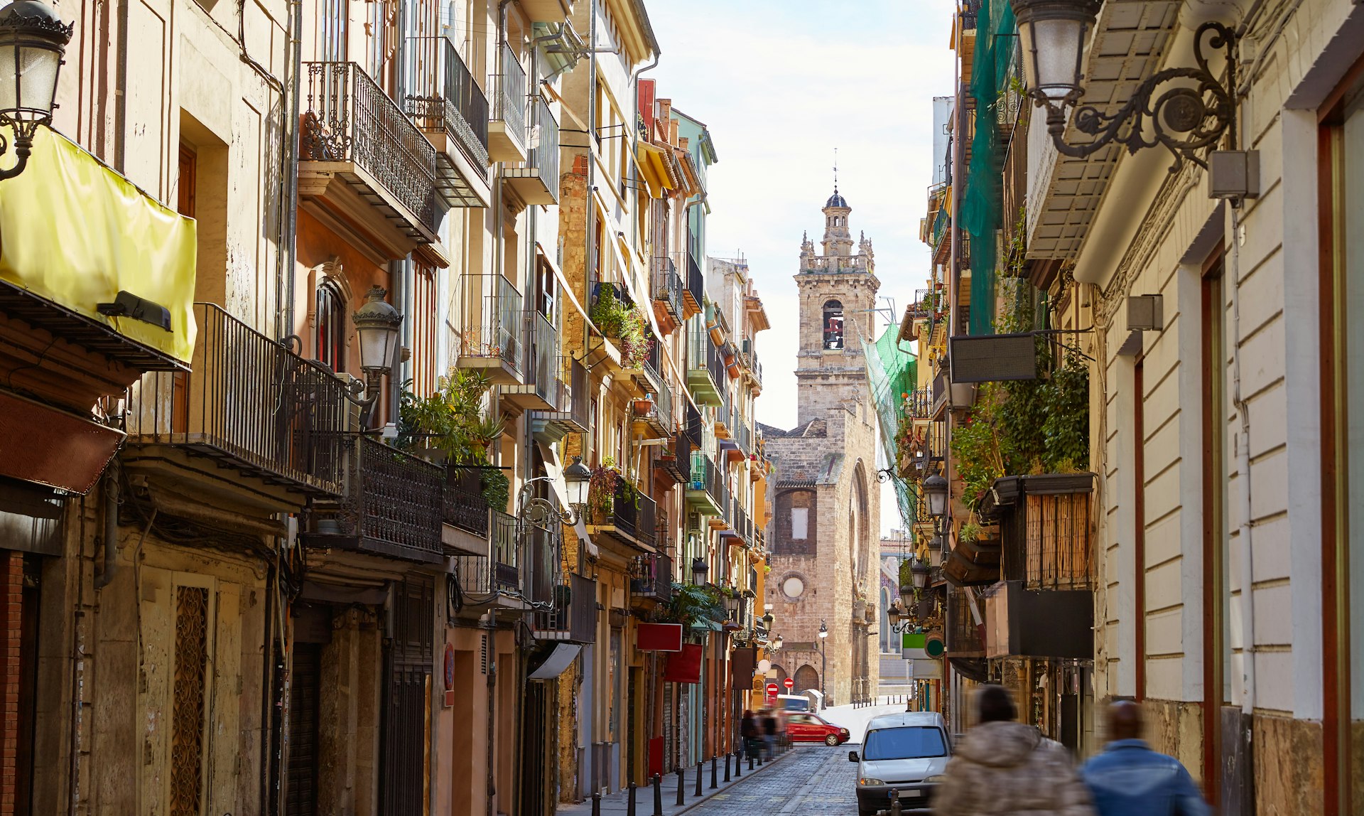 Buildings lining narrow Bolseria Street in Barrio del Carmen, with a church in the background and two blurry people in the foreground