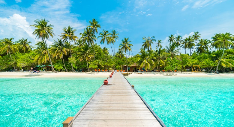 Maldives island resort with jetty, beach, sea and coconut palm trees on blue sky.